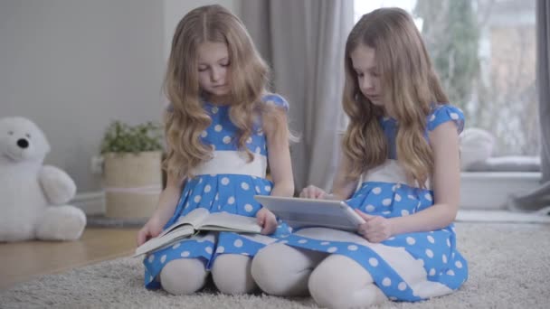 Brunette fille en utilisant la tablette comme sa sœur jumelle livre de lecture. Deux beaux enfants caucasiens dans des robes bleues similaires reposant à la maison. Mode de vie, loisir, passe temps . — Video