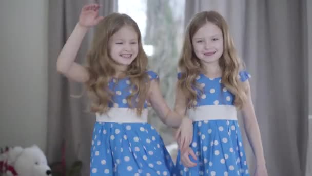 中间镜头是一对快乐的孪生姐妹在室内玩乐。快乐的黑发白人女孩，穿着类似的蓝色点缀裙，面带微笑。休闲、娱乐、生活方式、家庭. — 图库视频影像