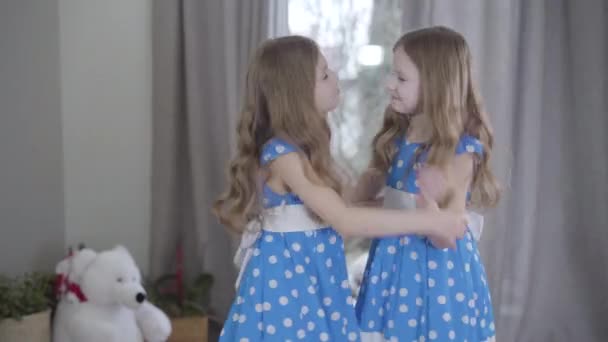 Pohled na dvě stejné sestry, které se baví doma. Radostná bělošská dvojčata v modrých tečkovaných šatech tančících uvnitř. Jednota, štěstí, životní styl. — Stock video