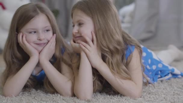 Close-up of happy Caucasian twin sisters looking at each other and turning to camera as lying and holding head with hands. Brunette girls with long wavy hair resting at home. Lifestyle, family. — Stock Video