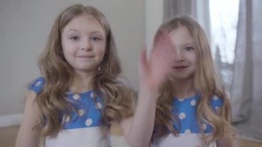 Positive twin sisters waving and sending air kisses to camera. Caucasian brunette girls in blue dotted dresses having video chat. Lifestyle, unity, joy, happiness.