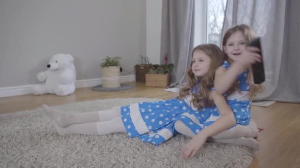 Two cute Caucasian girls sitting on soft carpet indoors with remote control. Twin sisters in blue dotted dresses watching TV at home. Leisure, unity, family. — ストック動画