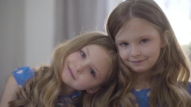Närbild av två små vita flickor i solstrålar tittar på kameran och ler. Ganska brunett barn sätta huvud på tvillingsystrar axel. Enhet, stöd, familj, livsstil. — Stockvideo
