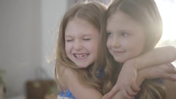 Close-up face of happy little pretty girl hugging twin sister and kissing her on cheek. Two children posing in sunlight indoors. Happiness, leisure, lifestyle, unity. — Stockvideo