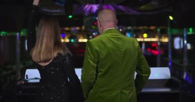 Back view of young attractive Caucasian woman and senior joyful man dancing on stage in night club. Cheerful female DJ mixing music as old male visitor having fun aside. Cinema 4k ProRes HQ.