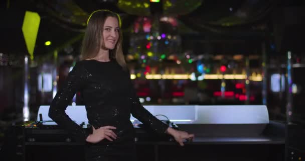 Portrait of gorgeous young Caucasian woman in black dress standing next to music controller, looking at camera and smiling. Professional beautiful female DJ posing in night club. Cinema 4k ProRes HQ. — Stockvideo