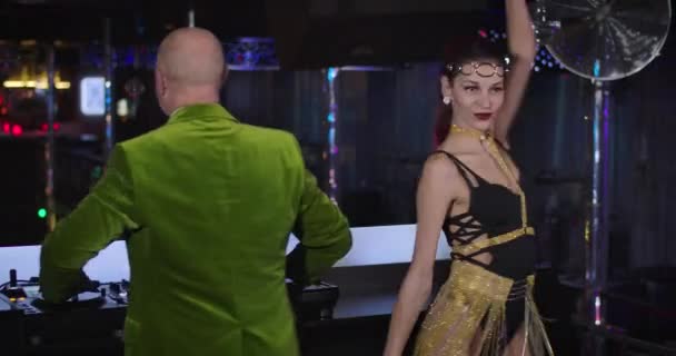 Seductive young Caucasian woman with tattoo on hand dancing on stage in night club next to elderly male DJ in green jacket mixing music on controller. Cinema 4k ProRes HQ. — Stockvideo