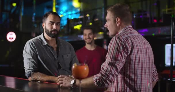 Young smiling Caucasian man coming to fellows in night club and greeting them. Three male friends standing in disco at bar counter and talking. Lifestyle, entertainment, resting. Cinema 4k ProRes HQ. — Stock Video