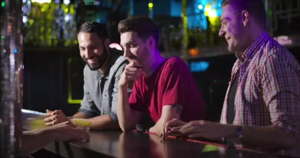 Three young men standing at bar counter and talking with bartender. Barkeeper giving short drinks to Middle Eastern and Caucasian fellows. Male friendship, resting, lifestyle. Cinema 4k ProRes HQ. — Stock Video