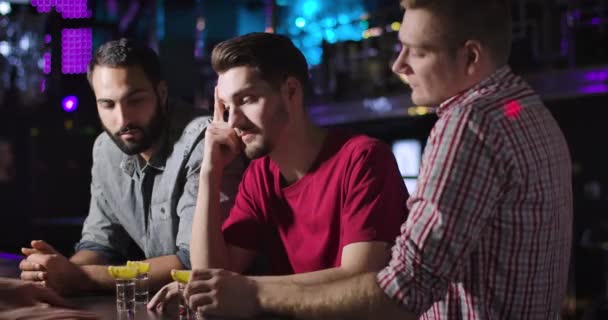 Primer plano de tres jóvenes hombres positivos tintineo vasos con bebidas cortas como de pie en el mostrador del bar en el club nocturno. Compañeros caucásicos y del Medio Oriente descansando con alcohol en la discoteca. Sede del cine 4k ProRes . — Vídeo de stock