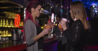 Two young beautiful women clinking glasses with cocktails and dancing next to bar counter in night club. Positive girls spending evening with alcohol in disco. Cinema 4k ProRes HQ.