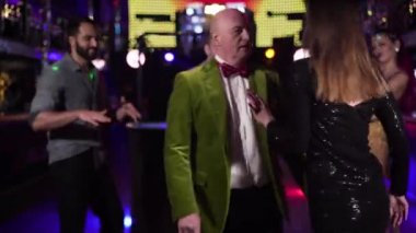 Young brunette Caucasian woman in black dress dancing around elderly man in night club. Senior man in green jacket spending evening with younger lady. Lifestyle, entertainment.