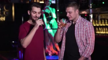 Two young Caucasian men clinking glasses and drinking alcohol in night club. Male friends resting with PJ dancing at the background. Lifestyle, leisure, joy.