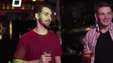 Close-up of two handsome Caucasian men talking and drinking alcohol in night club. Young positive guys resting in disco as go go dancer moving at the background. Entertainment,. leisure, lifestyle.