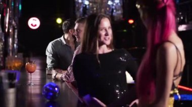 Focus changes from Middle Eastern and Caucasian men talking at the background to young beautiful female friends chatting at the foreground. Multi ethnic people resting in disco.