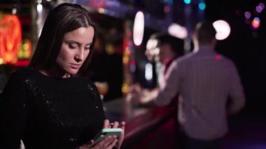 Portrait of adult beautiful Caucasian woman in black dress standing at bar counter in night club and using smartphone. Attractive girl spending evening in disco alone. Lifestyle, leisure, loneliness.