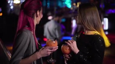 Close-up of two young pretty Caucasian girls looking back at visitors in night club, talking and laughing. Joyful women spending evening with cocktails in disco. Leisure, lifestyle, joy.