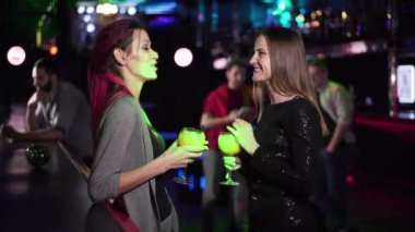 Two positive Caucasian girls standing with cocktails at bar counter, talking and waving to men sitting at the background. Beautiful women spending evening in night club. Entertainment, lifestyle.