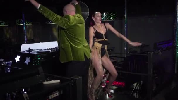 Elderly Caucasian man and seductive young woman dancing on stage next to music controller in night club. Portrait of positive DJ and go go dancer in disco. Lifestyle, profession, entertainment. — Stok video