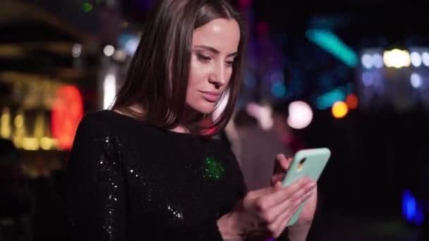 Close-up of confident young Caucasian woman using smartphone in night club. Gorgeous girl in black dress typing on screen as resting in disco in the evening. Leisure, lifestyle, relaxation. — ストック動画