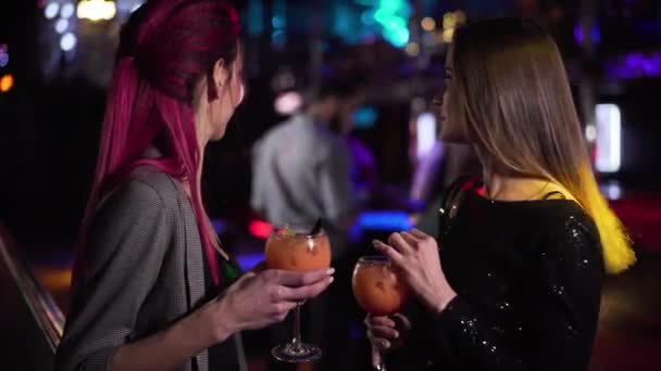 Close-up of two young pretty Caucasian girls looking back at visitors in night club, talking and laughing. Joyful women spending evening with cocktails in disco. Leisure, lifestyle, joy. — Stok video