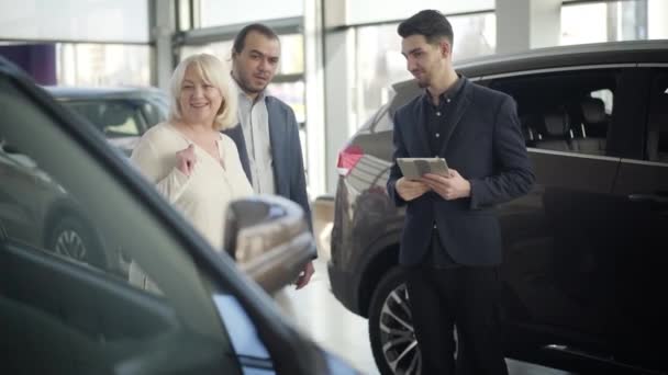 Mulher caucasiana sênior e homem discutindo novo automóvel com comerciante no showroom. Clientes positivos confiantes escolhendo carro na concessionária. Negócios, estilo de vida, sucesso, indústria . — Vídeo de Stock