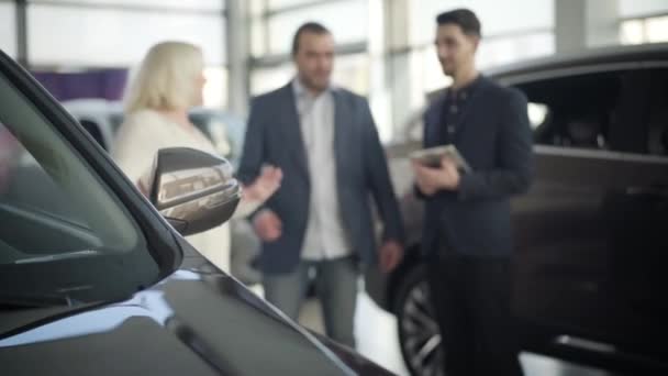 Nahaufnahme eines neuen schwarzen Autos, das im Showroom steht, während sich im Hintergrund drei verschwommene Personen unterhalten. Erwachsenes Paar im Gespräch mit männlichen Händlern im Autohaus. Unternehmen, Industrie, Lebensstil. — Stockvideo