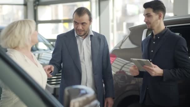 Adult confident Caucasian businessman shaking traders hand and showing wife new automobile in showroom. Successful brunette man purchasing vehicle for positive blond woman in dealership. — Stock video