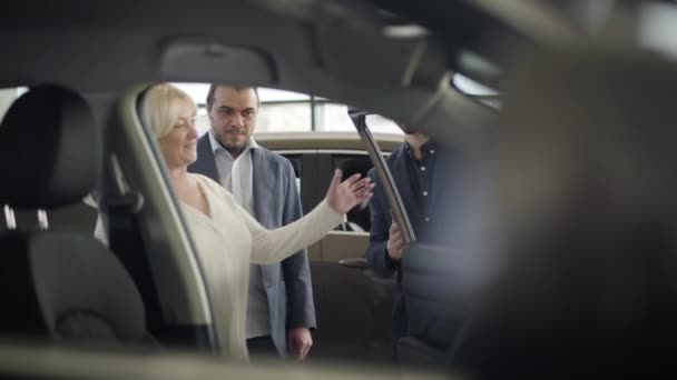 Молодой кавказский автодилер открывает дверь для взрослого блондинистого клиента, старшая женщина сидит на водительском сидении. Счастливая пара выбирает автомобиль в дилерском центре. Изменение фокуса от фона к фронту . — стоковое видео