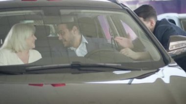Araba kuaföründe öpüşen ve gülümseyen yaşlı beyaz bir çiftin portresi. Yeni otomobilin mutlu sahipleri içeride oturuyor ve arabanın yanında duran erkek tüccarla konuşuyor. Başarı, neşe, yaşam tarzı.