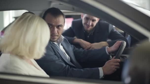 Focus changes from face of confident Caucasian businessman on drivers seat to happy blond mature woman sitting on passenger seat and making victory gesture. Happy wife getting gift from husband. — Stock video