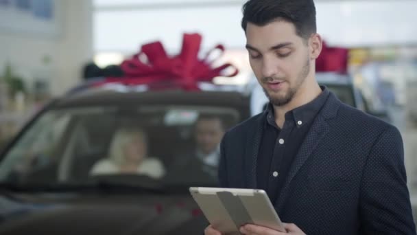 Close-up portrait of young handsome brunette Caucasian man with brown eyes looking back at couple of customers, turning to camera and showing thumb up. Confident car dealer standing in dealership. — Stock video