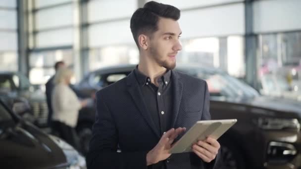 Young handsome brown-eyed Caucasian man standing in car dealership with tablet, looking at camera and smiling. Confident brunette dealer posing at workplace in showroom. Job, occupation, lifestyle. — Stock Video