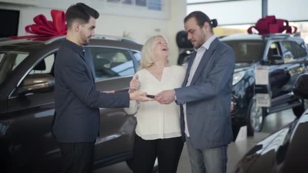 Young professional car dealer giving keys from new vehicle to senior couple, grateful blond woman kissing husband on cheek, man shaking traders hand. Businessman buying car for wife in showroom. — Stock video