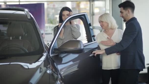 Handsome young Caucasian trader opening car door for mature blond woman choosing new automobile with beautiful brunette daughter. Successful businesswoman buying gift for mother in dealership. — Stock video