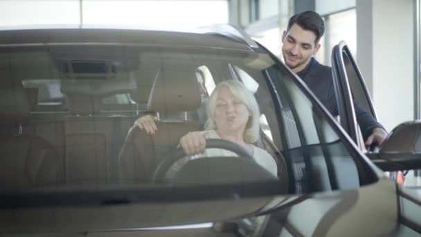 Jonge glimlachende autodealer die klanten raadpleegt die in de auto zitten. Rijpe blonde vrouw en jonge brunette dochter kiezen voertuig in de dealerovereenkomst. Succes, rijkdom, levensstijl, bedrijfsleven. — Stockvideo