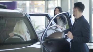 Genç, kendine güvenen beyaz iş kadını olgun anne ve tüccar için yeni bir otomobil satın almayı tartışıyor. Yaşlı sarışın kadın arabada oturuyor, gülümseyen esmer kadın araba satıcısıyla konuşuyor..