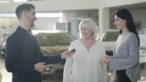 Jovem comerciante caucasiano dando chaves de carro para mulher madura, mãe passando-os para menina morena animado. Senhora loira feliz comprando presente para a filha na concessionária. Alegria, felicidade, riqueza, estilo de vida . — Vídeo de Stock