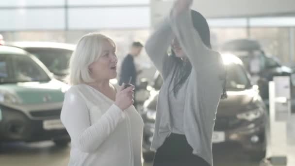 Happy young brunette woman taking car keys from positive mature mother, dancing and hugging parent. Excited Caucasian daughter receiving gift in dealership on sunny day. Joy, wealth, success. — Stock video