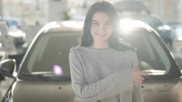 Portrait of beautiful brunette Caucasian woman with green eyes standing in sunlight in car dealership and winking at camera. Happy joyful girl buying automobile in showroom. Joy, happiness, wealth. — Stock Video