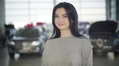 Siyah saçlı, yeşil gözlü, kameraya bakıp gülümseyen beyaz bir kadının portresi. Araba galerisinde poz veren güzel bir bayan. İş, yaşam tarzı, mutluluk.