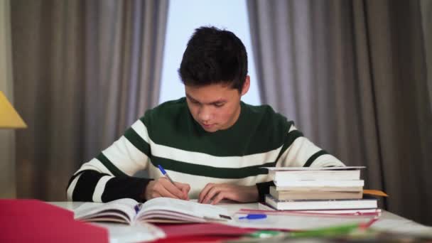 Portret van een gelukkige Aziatische jongen die binnen studeert. Leuke tiener student zit aan de tafel met boeken en glimlachen. Lifestyle, onderwijs, intelligentie. — Stockvideo