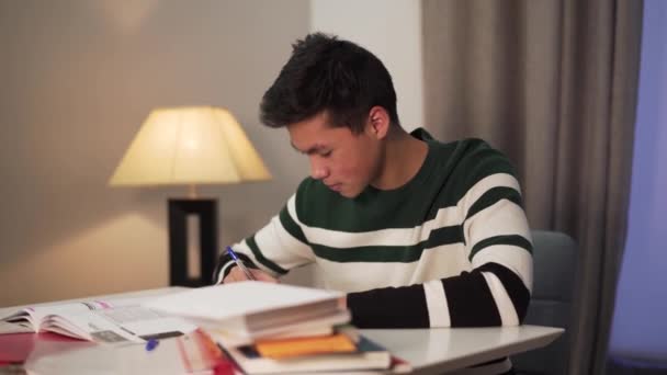 Вид сбоку взволнованный азиатский студент делает домашнее задание в помещении. Мальчик-подросток сидит за столом с книгами и улыбается. Образование, интеллект, образ жизни . — стоковое видео