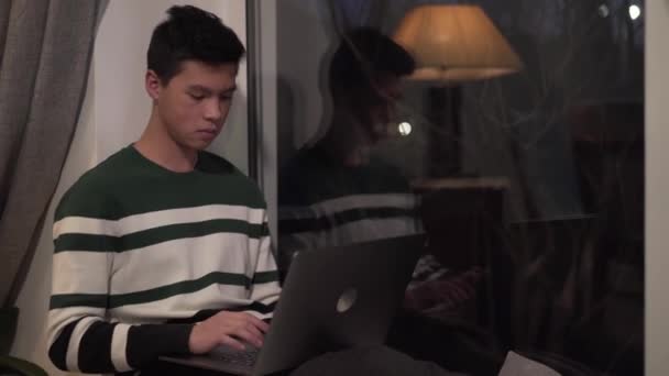 Portrait of young Asian boy typing on laptop and looking out the window in the evening. Handsome college student studying indoors. Ideas, intelligence, online education. — Stock Video