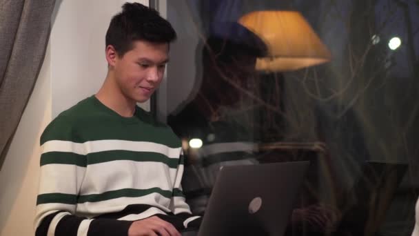 Feliz adolescente asiático sentado en el alféizar de la ventana y el uso de la computadora portátil. Hermoso estudiante universitario escribiendo en el teclado y sonriendo. Internet, redes sociales, tecnologías modernas . — Vídeos de Stock