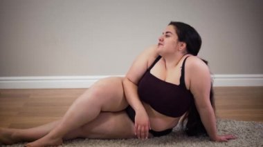 Yere oturmuş gülümseyen, iç çamaşırlı, kendine güvenen beyaz kadın portresi. İçerde poz veren genç obez kız. Aşırı kilo problemi, obezite, yaşam tarzı.