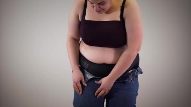 Portrait of positive fat Caucasian girl putting in jeans. Obese young woman jumping trying to pull up trousers. Cellulite, obesity, fatness. — Stock Video