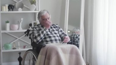 Kafkasyalı, beyaz saçlı, tekerlekli sandalyede oturan ve şiddetli baş ağrısından muzdarip emeklinin portresi. Yaşlı ve felçli bir adam bütün gününü evde yalnız geçiriyor. Engellilik, yaşam tarzı, yaşlanma.