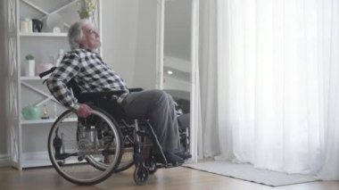 Kafkasyalı, üzgün, engelli bir adamın portresi. Tekerlekli sandalyeyi pencereden indiriyor ve yuvarlanıyor. Depresyondaki erkek emekli, içeride hareket etmek için ekipman kullanıyor. Yalnızlık, sakatlık, yaşlanma..
