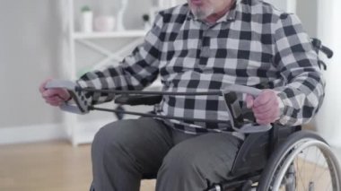 Tekerlekli sandalyede elastik bant takan tanınmayan beyaz bir adam. Evde egzersiz yapan yaşlı, engelli bir erkek. Fizik tedavi, sağlık hizmetleri, engellilik.